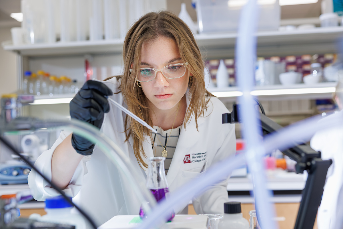 Texas A&M medical student in a white lab coat using a pipette to transfer liquid into a flask in a research laboratory.